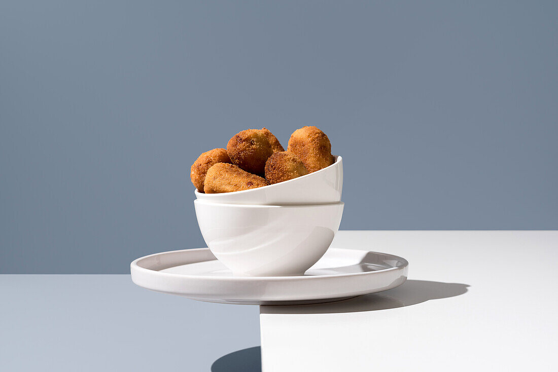 A white bowl on a saucer, filled with croquettes, presented against a grey-blue background, creating a modern and minimalist food aesthetic