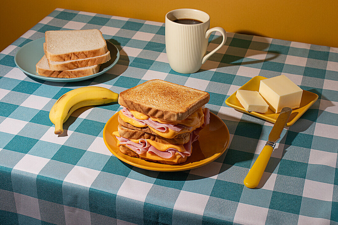 Ein Sandwich auf einem gelben Teller wird zusammen mit einer Tasse Kaffee, Butter auf einem kleinen Teller und einer frischen Banane auf einer blau-weiß karierten blauen Tischdecke serviert.