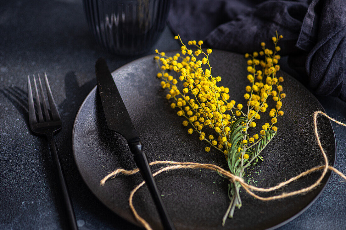 From above a sophisticated black plate setting accented by a fork, knife, and a bright spray of yellow mimosa flowers bundled with twine.