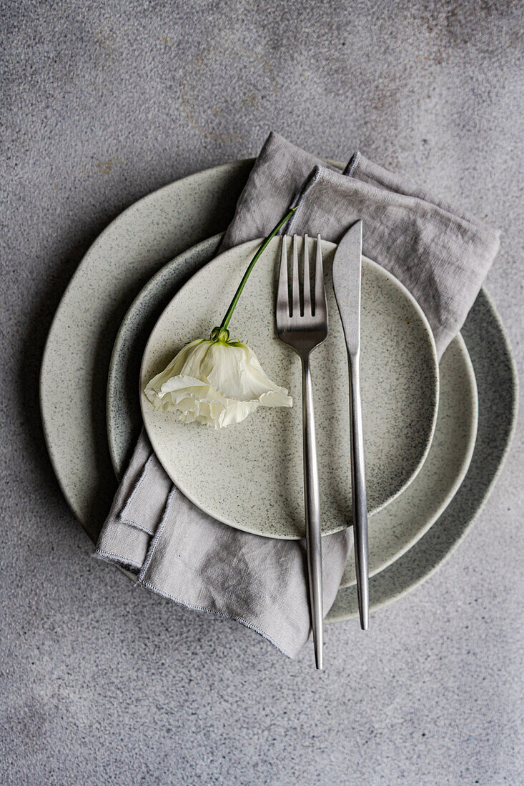 Top view a refined table setting featuring layered ceramic plates and a soft gray napkin, accentuated by a delicate godetia flower lying gently across the setup