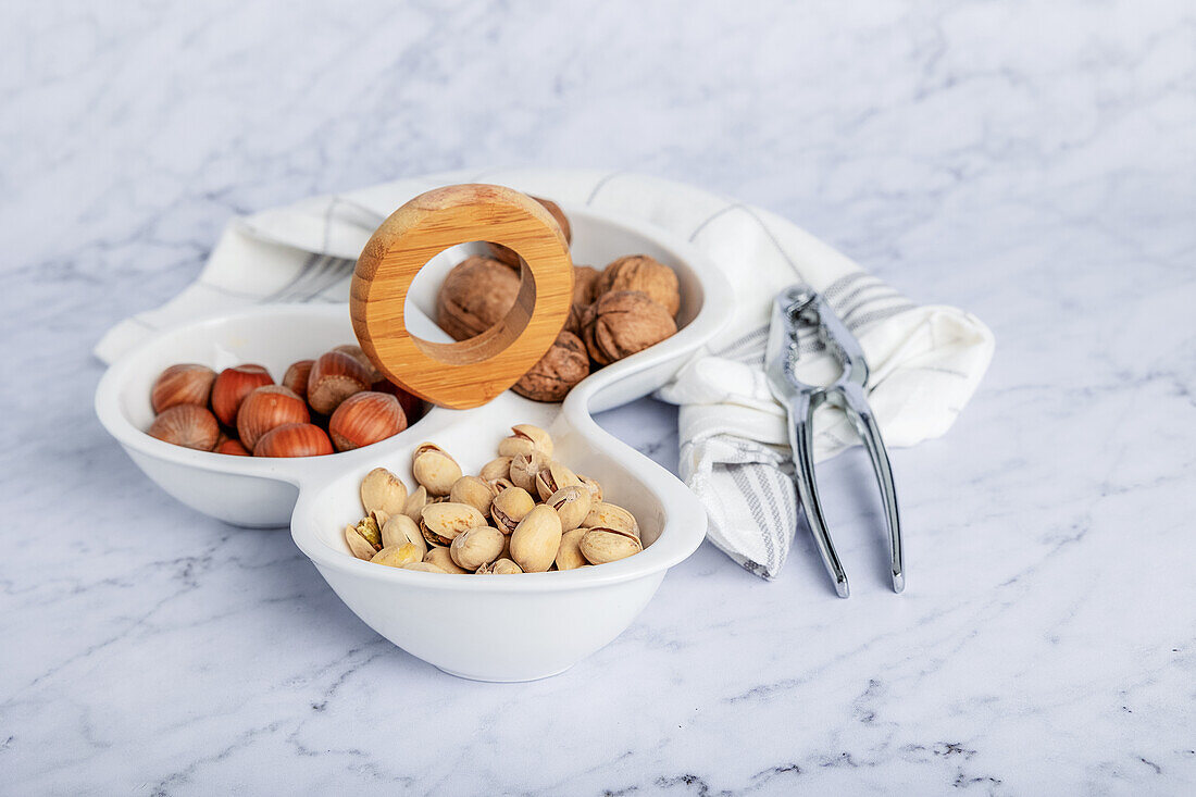 From above marble table setting showcasing a wholesome selection of nuts including walnuts, pistachios, and hazelnuts in white bowls. A wooden nutcracker adds a functional touch to the scene, emphasizing a healthy snack option.