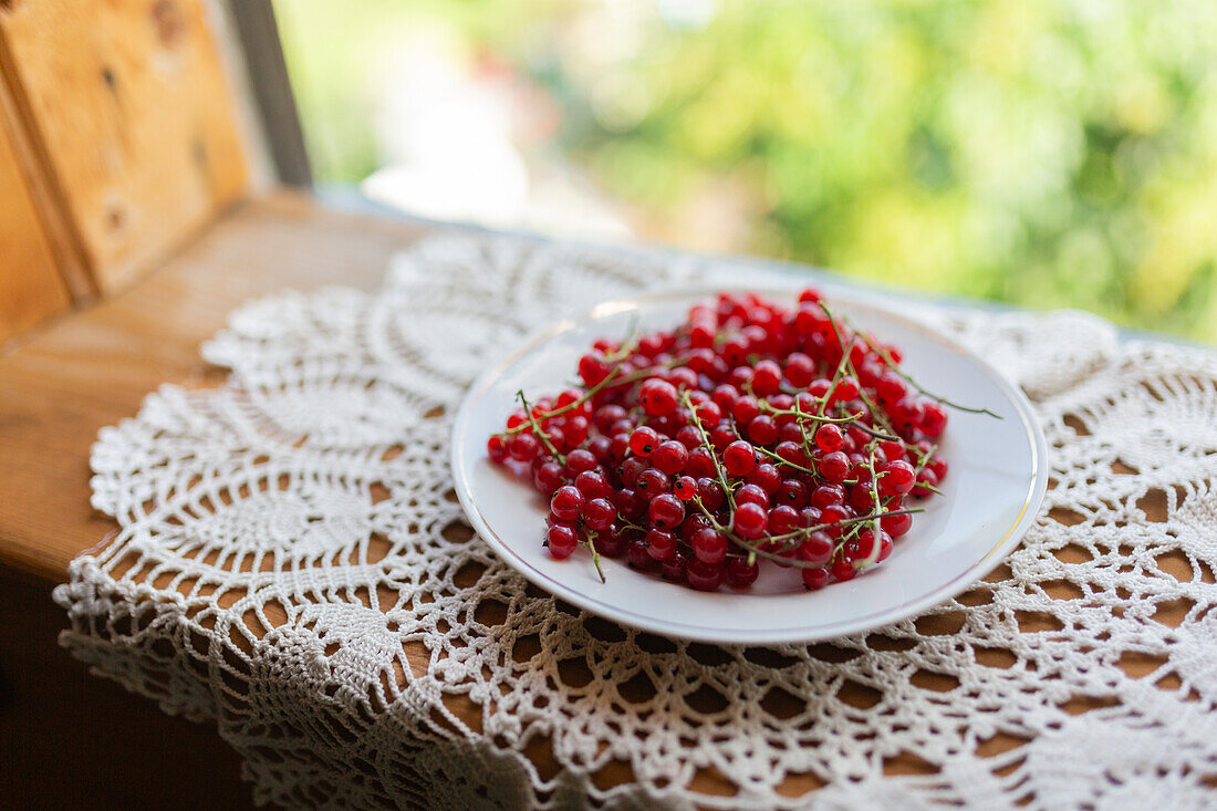 Frische rote Johannisbeeren auf einem weißen Teller auf einer kunstvollen Spitzentischdecke Das natürliche Tageslicht unterstreicht die satte, rote Farbe der Beeren und sorgt für eine herrliche Sommeratmosphäre