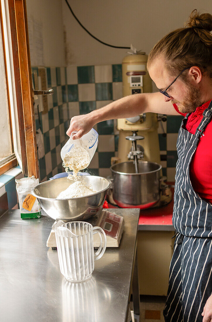 A skilled chef, wearing a striped apron, prepares pizza dough by pouring flour into a metal mixing bowl in a kitchen equipped with vintage machinery