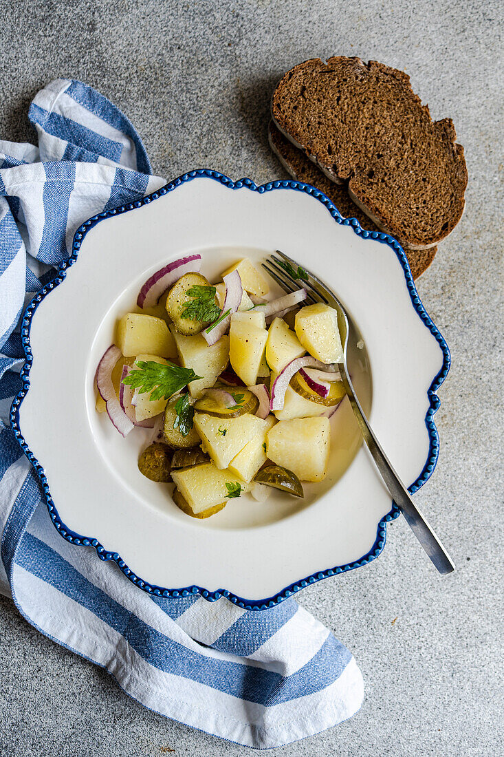 A classic variation of German potato salad, featuring diced boiled potatoes, crisp red onions, and tangy slices of fermented cucumber served in a decorative white and blue rimmed bowl with a side of dark rye bread