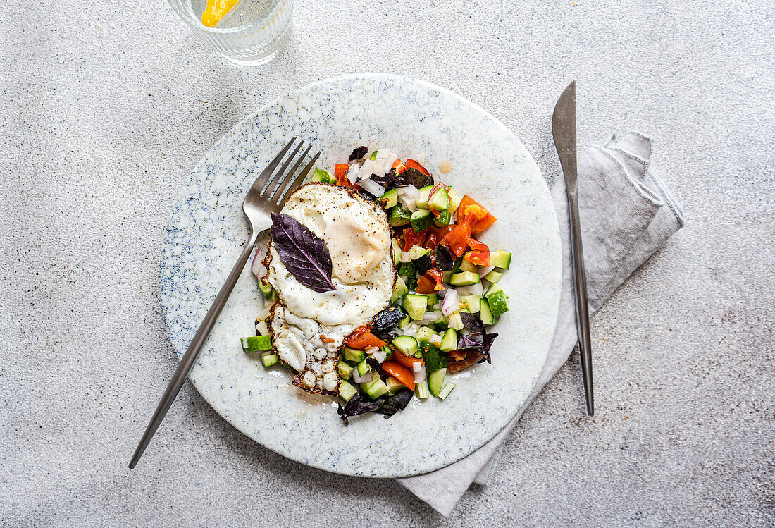 Ein nahrhaftes, ketofreundliches Mittagessen mit einem frisch gekochten Ei auf einem bunten gemischten Salat mit Gurken, Tomaten und lila Basilikum, serviert auf einem marmorierten Teller mit elegantem Silberbesteck