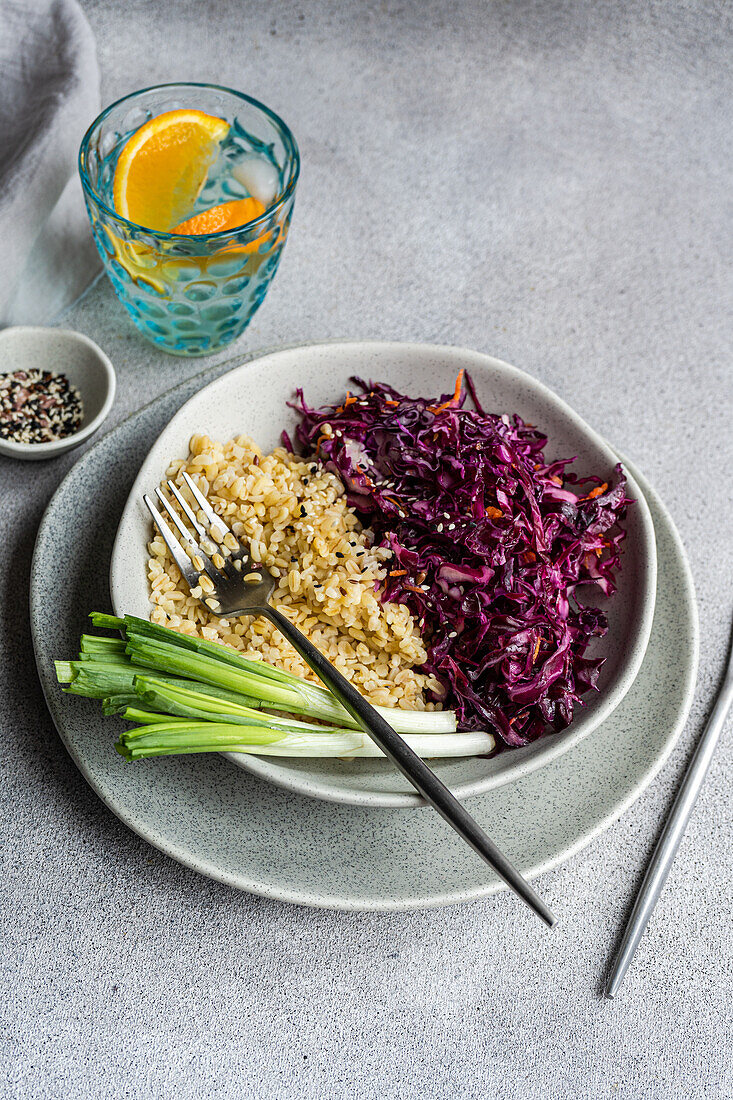 Eine nahrhafte Mahlzeit mit lebhaftem rohem Rotkohl- und Karottensalat und lockerem gekochtem Bulgur, präsentiert auf einem grauen Keramikteller mit frischen grünen Zwiebeln