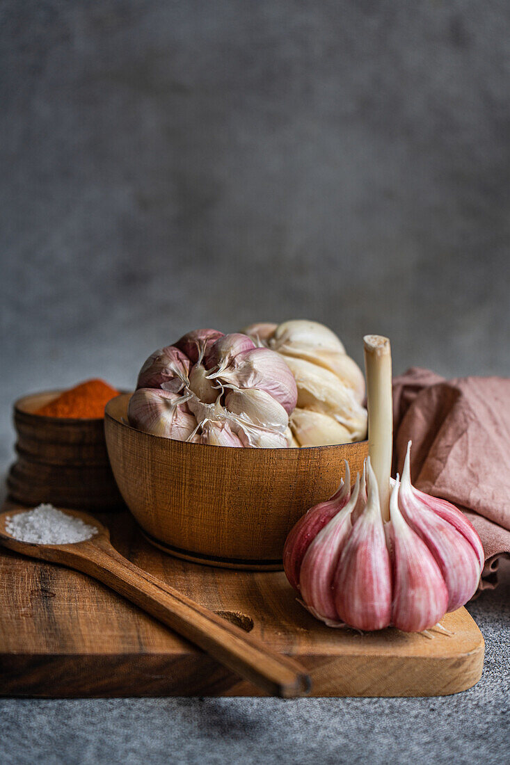 A carefully arranged set of cooking ingredients featuring fresh garlic bulbs in a wooden bowl, accompanied by spices and a salt spoon, set against a textured gray backdrop