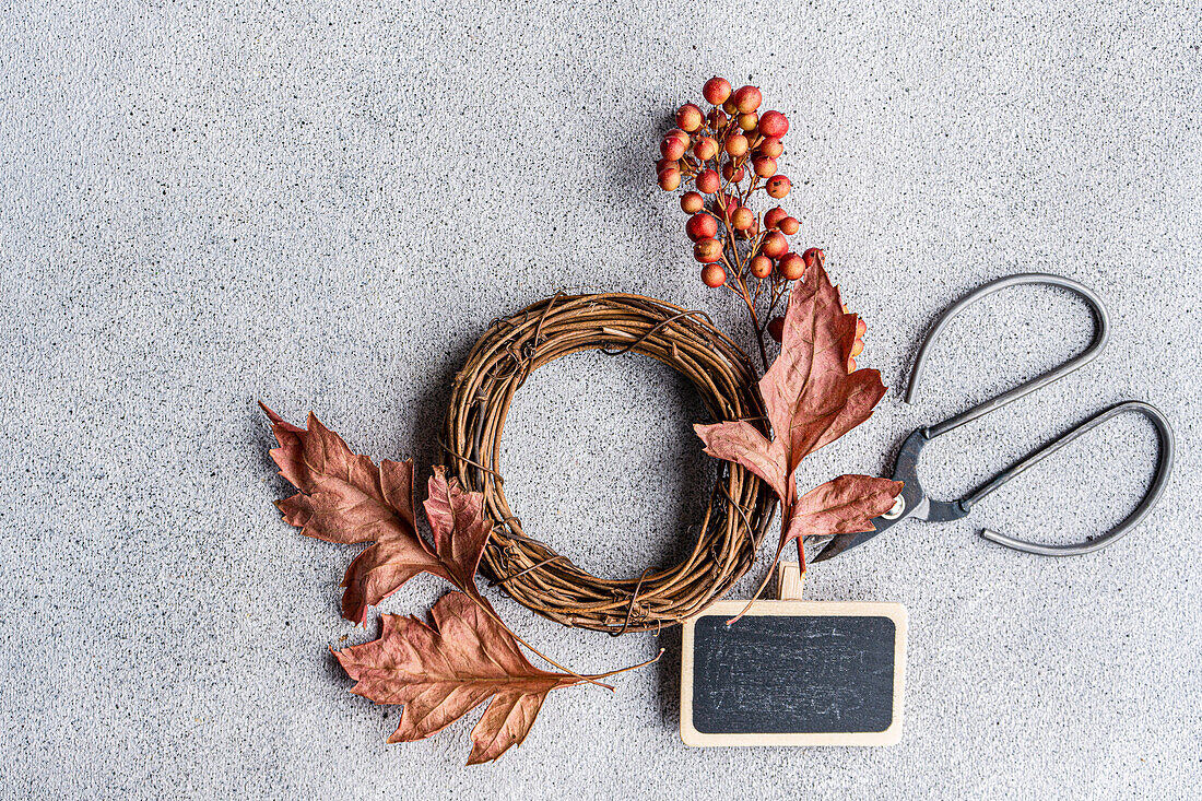 Creative flat lay of autumn wreath making supplies including dried leaves, a grapevine wreath, red berries, and shears with a blank chalkboard, on a textured concrete background