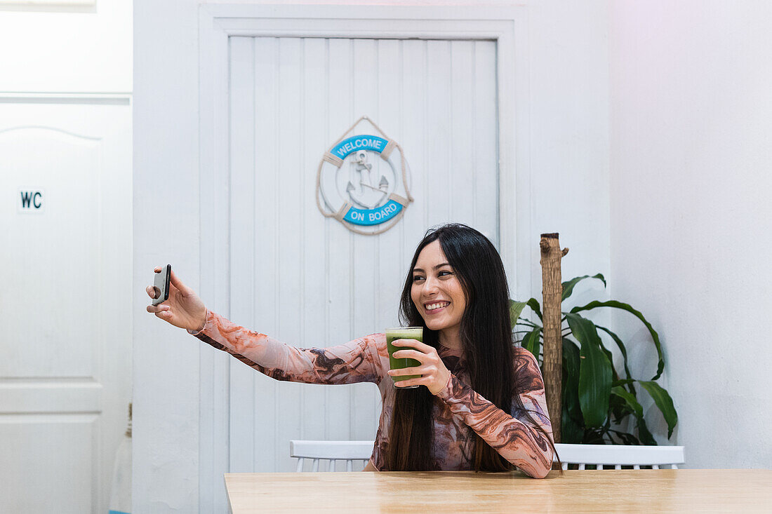 Positive female sitting at table with smoothie while taking selfie with mobile phone while chilling at weekend in bar