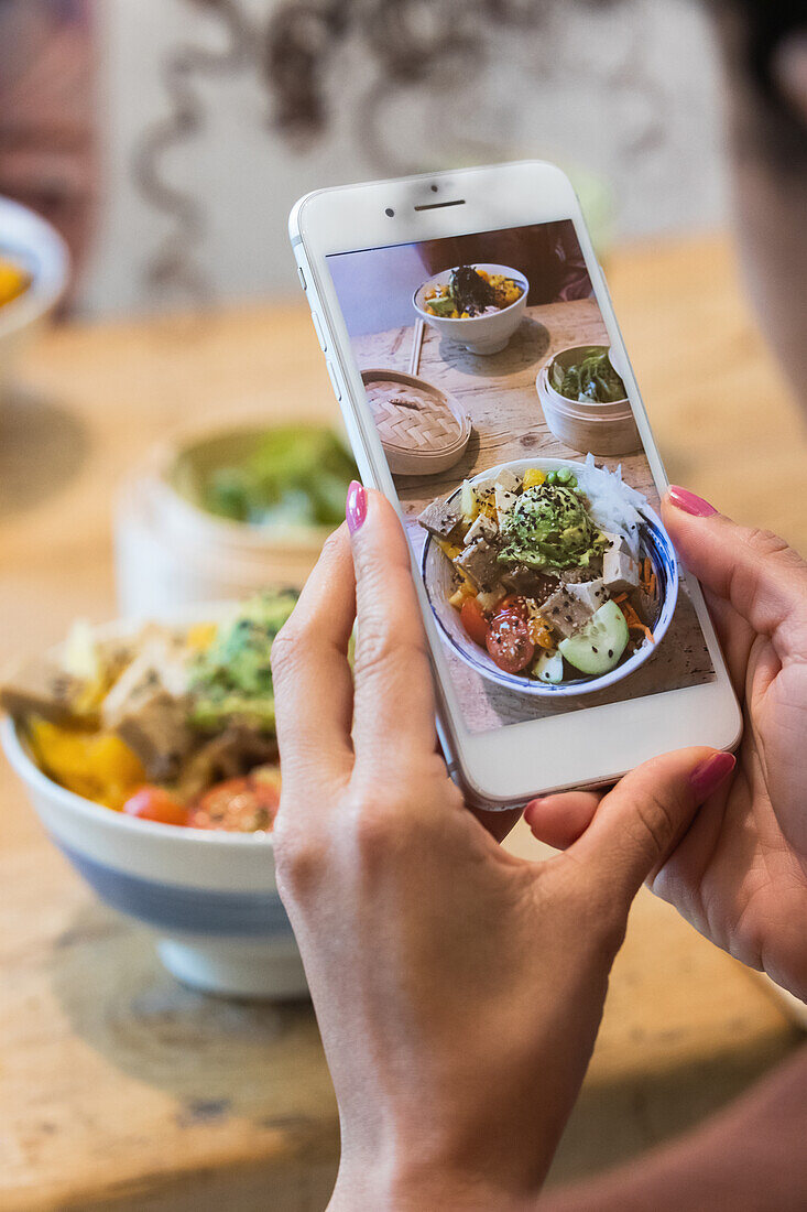 Abgeschnittene, nicht erkennbare Frau, die mit ihrem Smartphone ein Foto von einem Poke-Gericht macht, während sie mit einer Freundin ein Wochenende im Restaurant verbringt