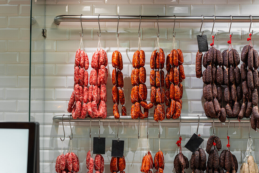 A variety of chorizos and sausages are displayed hanging on metal hooks against a tiled wall in a deli