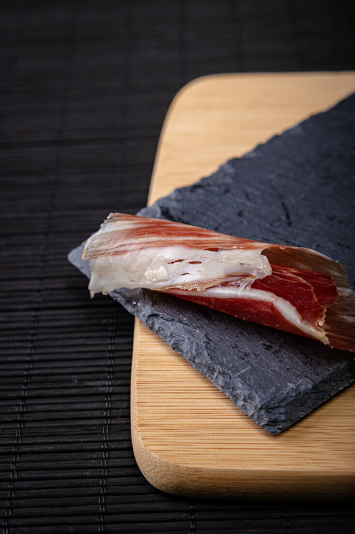 A close-up image showcasing thin slices of Serrano ham artistically placed on a slate board atop a wooden surface, exemplifying a quintessential Spanish delicacy