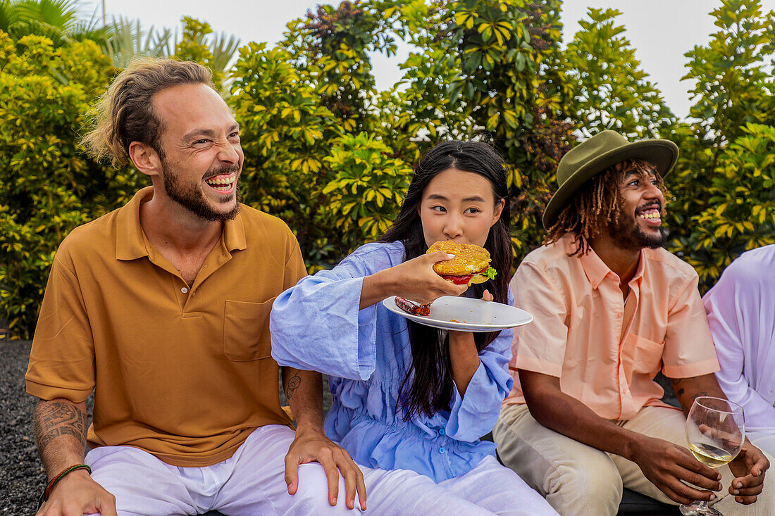 A diverse group of friends shares laughter and food at an outdoor barbecue. An Asian woman eats a burger, a Caucasian man laughs, and an African American man smiles, all enjoying the moment.