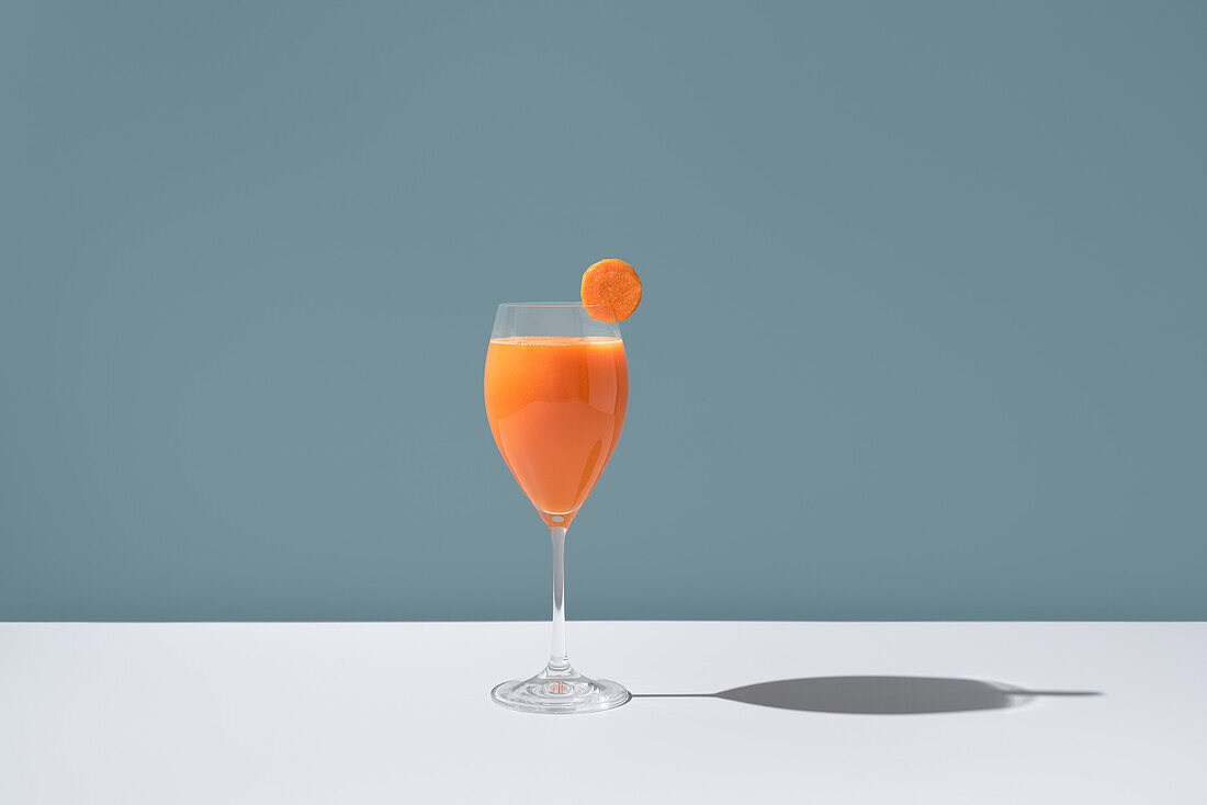 An elegant glass filled with vibrant orange carrot juice, garnished with a fresh carrot slice placed on the rim, set against a soft blue background.