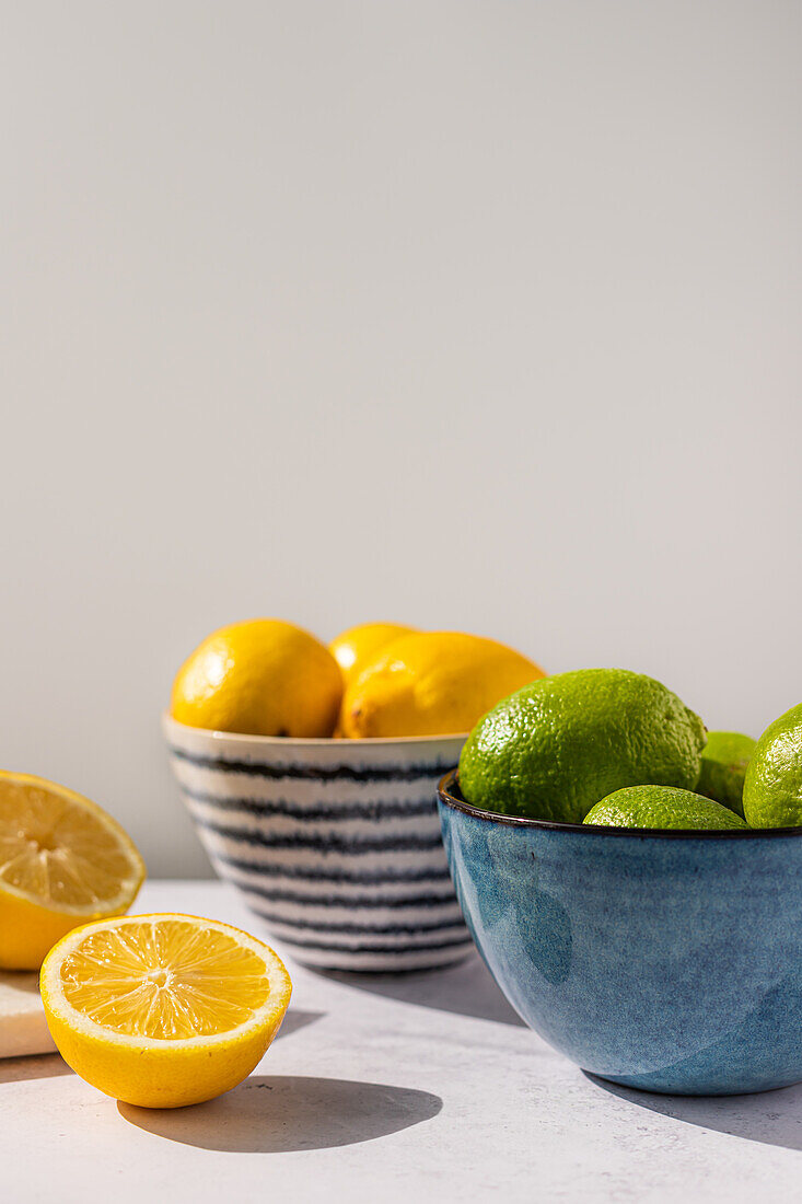 Bowls of vibrant yellow lemons and green limes presented on a light surface, with a half-cut lemon in the foreground