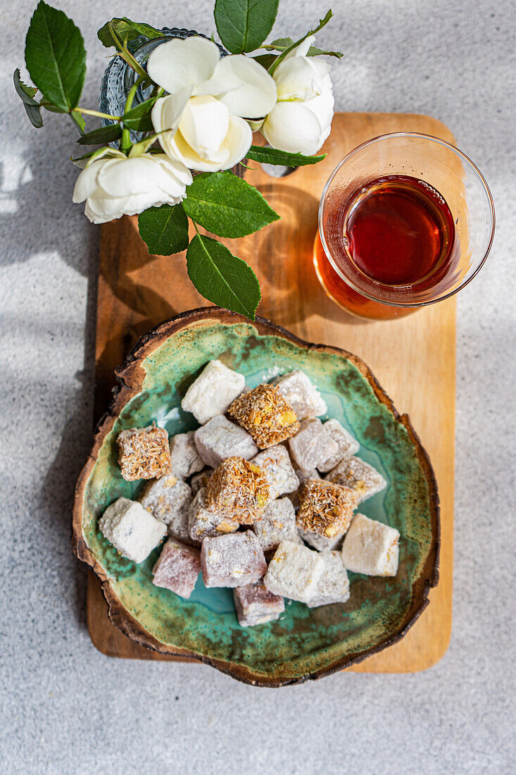 A mix of dusted Turkish delights on a ceramic plate with a glass of black tea, accompanied by a vase of white roses, all presented on a wooden board