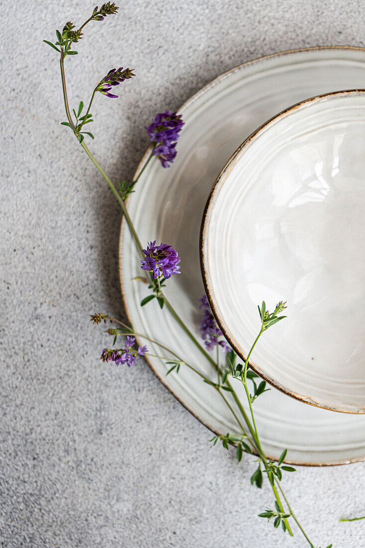 Top view of a minimalist table setting featuring delicate purple wildflowers alongside rustic ceramic plates on a textured surface, capturing the essence of summer's simplicity.