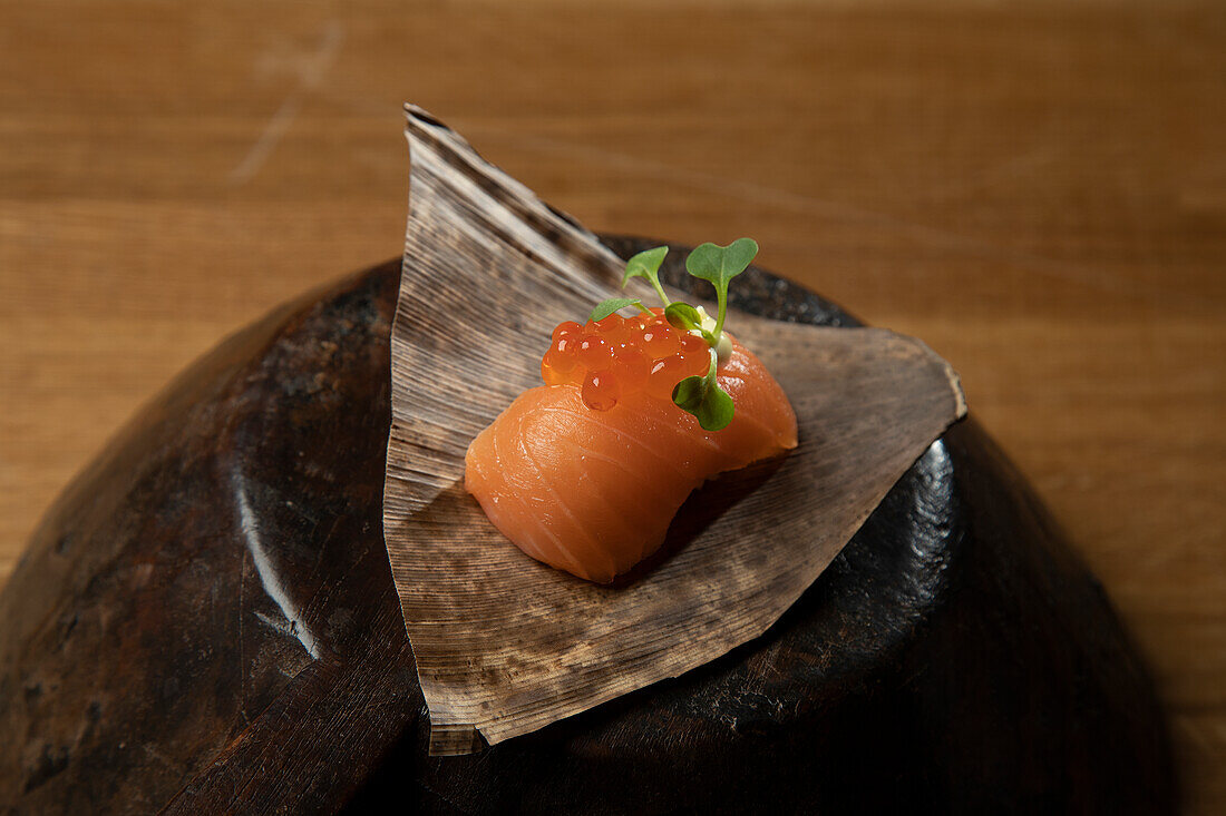 A beautifully presented salmon sashimi, delicately placed on a traditional leaf, garnished with vibrant salmon roe and a sprig of fresh greens, resting on a dark, rustic wooden bowl