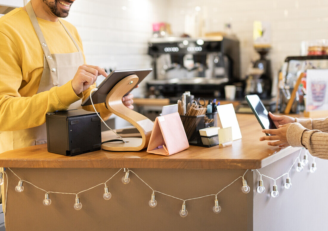 Ein fröhlicher Barista in einem gelben Pullover nimmt eine kontaktlose Zahlung von einem Kunden an einem gemütlichen Café entgegen.