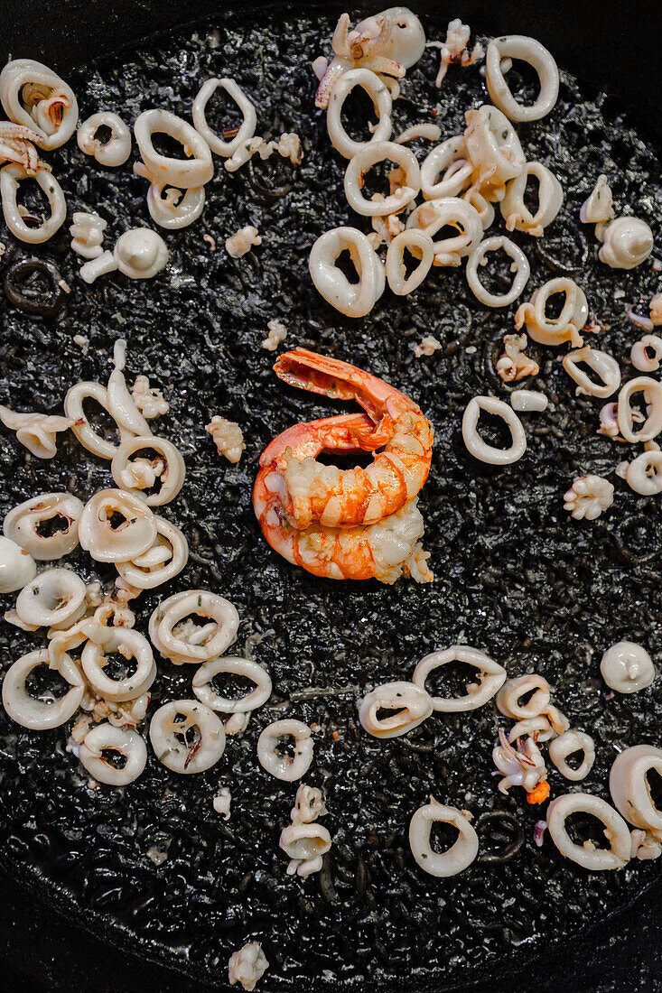 Draufsicht auf eine traditionelle Paella mit schwarzem Reis, gekocht mit Tintenfischtinte, Baby-Tintenfischen und Garnelen, die den valencianischen Stil mit einem perfekten Socarrat demonstriert.