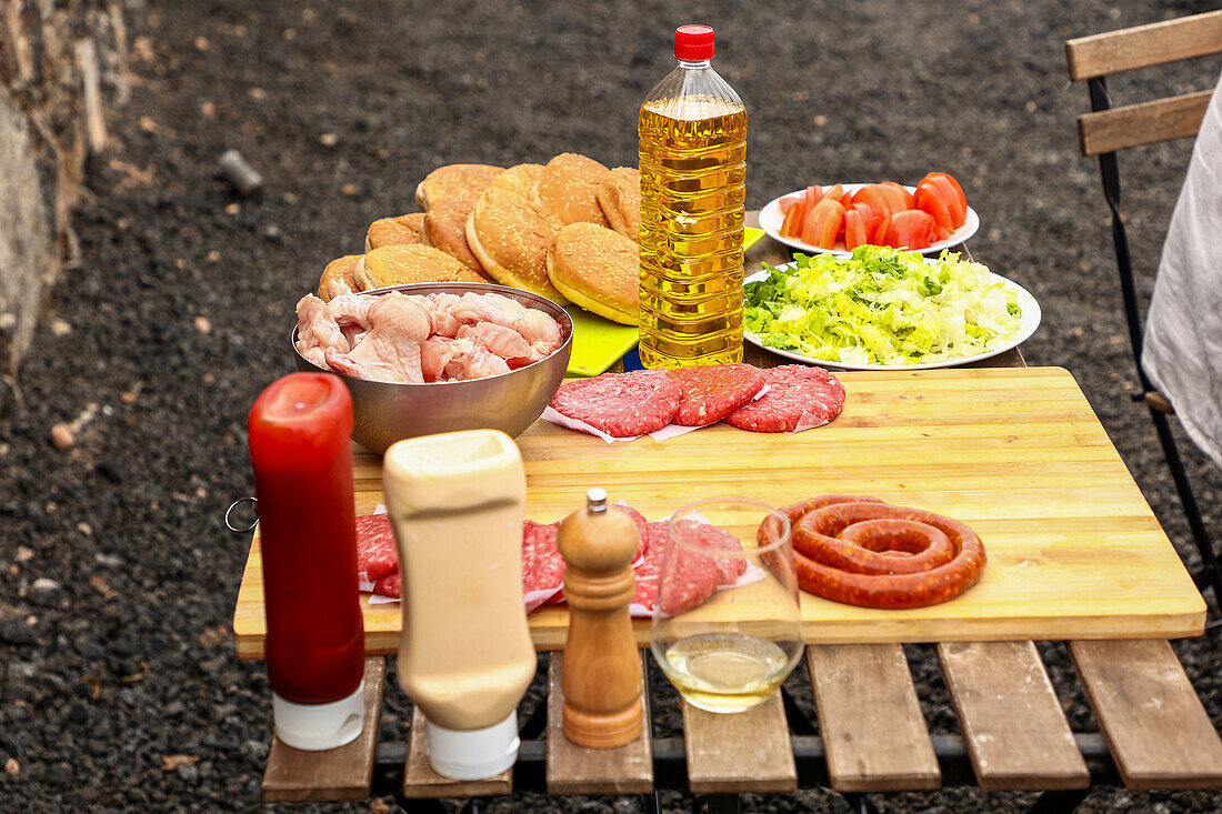 A ready-to-grill outdoor barbecue selection featuring raw meats, buns, and salads on a wooden board, complete with condiments and a bottle of oil.