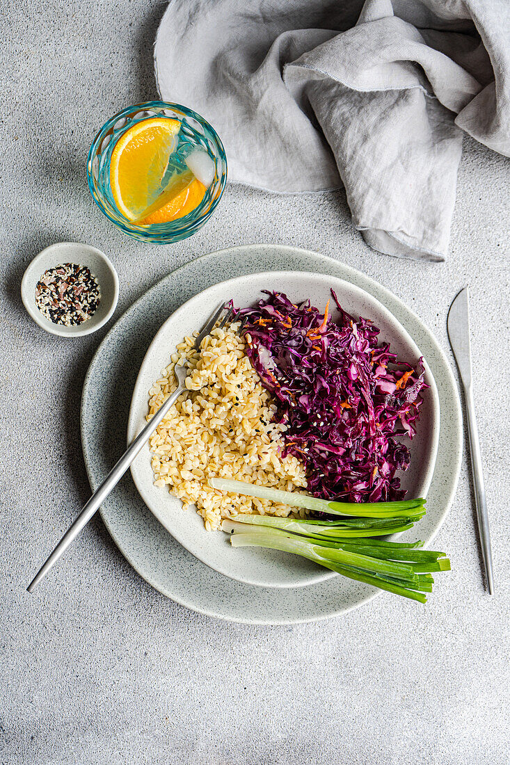 A nutritious and vibrant lunch setup featuring a raw red cabbage and carrot salad alongside steamed bulgur, served with green onion and a citrus water