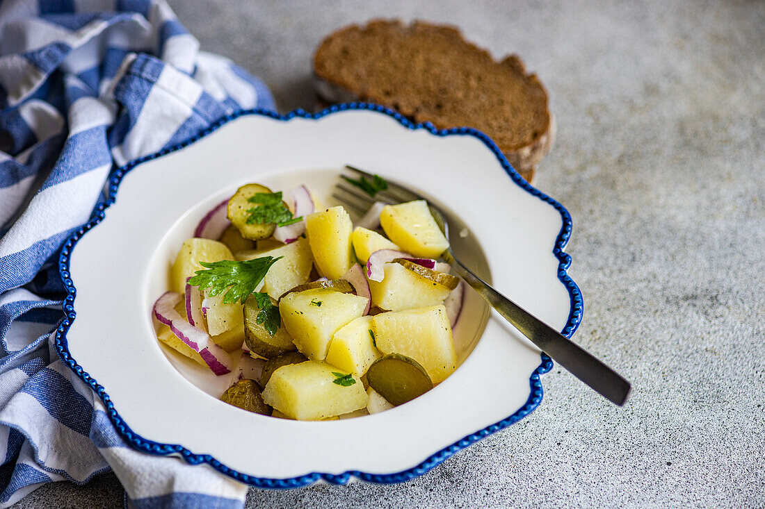 Traditional German potato salad featuring boiled potatoes mixed with slices of red onion and fermented cucumber, presented in a decorative white and blue bowl, accompanied by slices of rye bread