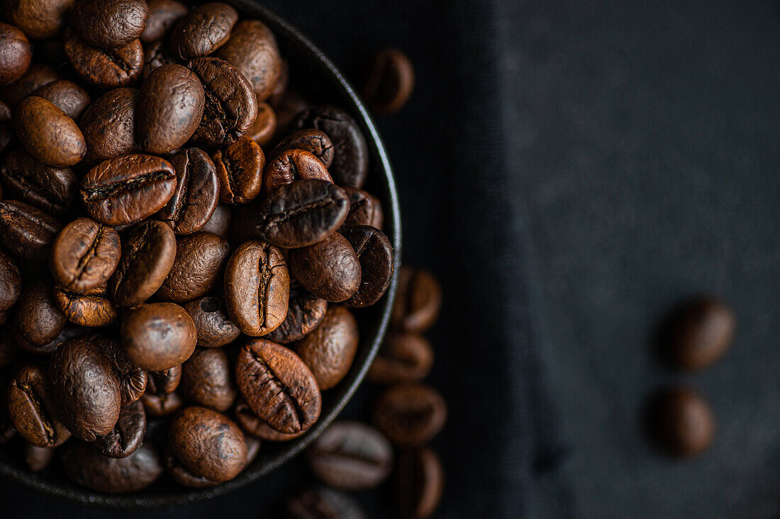 Draufsicht auf eine dunkle Schale mit glänzenden, frischen Kaffeebohnen vor einem dunkel strukturierten Hintergrund, der die reichhaltigen Strukturen und tiefbraunen Farben hervorhebt.