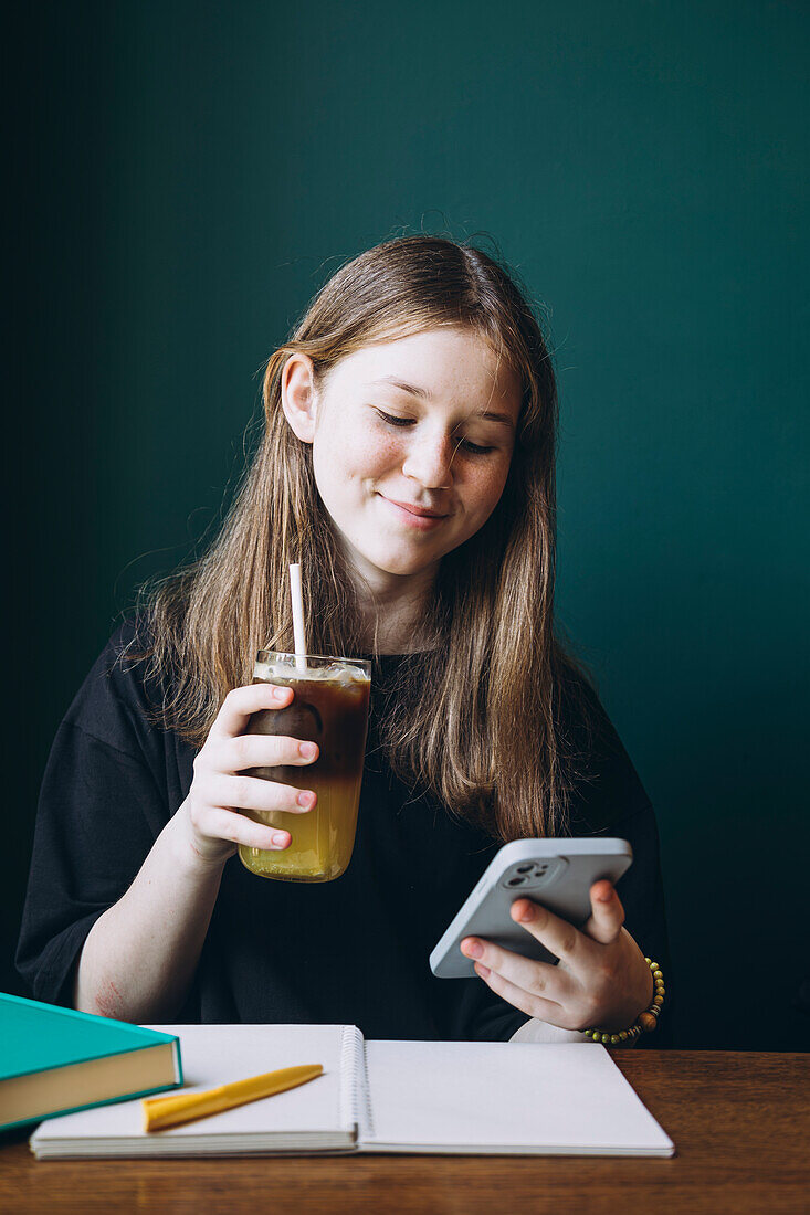 A young happy female student enjoys a refreshing iced tea while browsing her smartphone. She sits at a wooden table with school supplies, looking down at her device, immersed in its content.