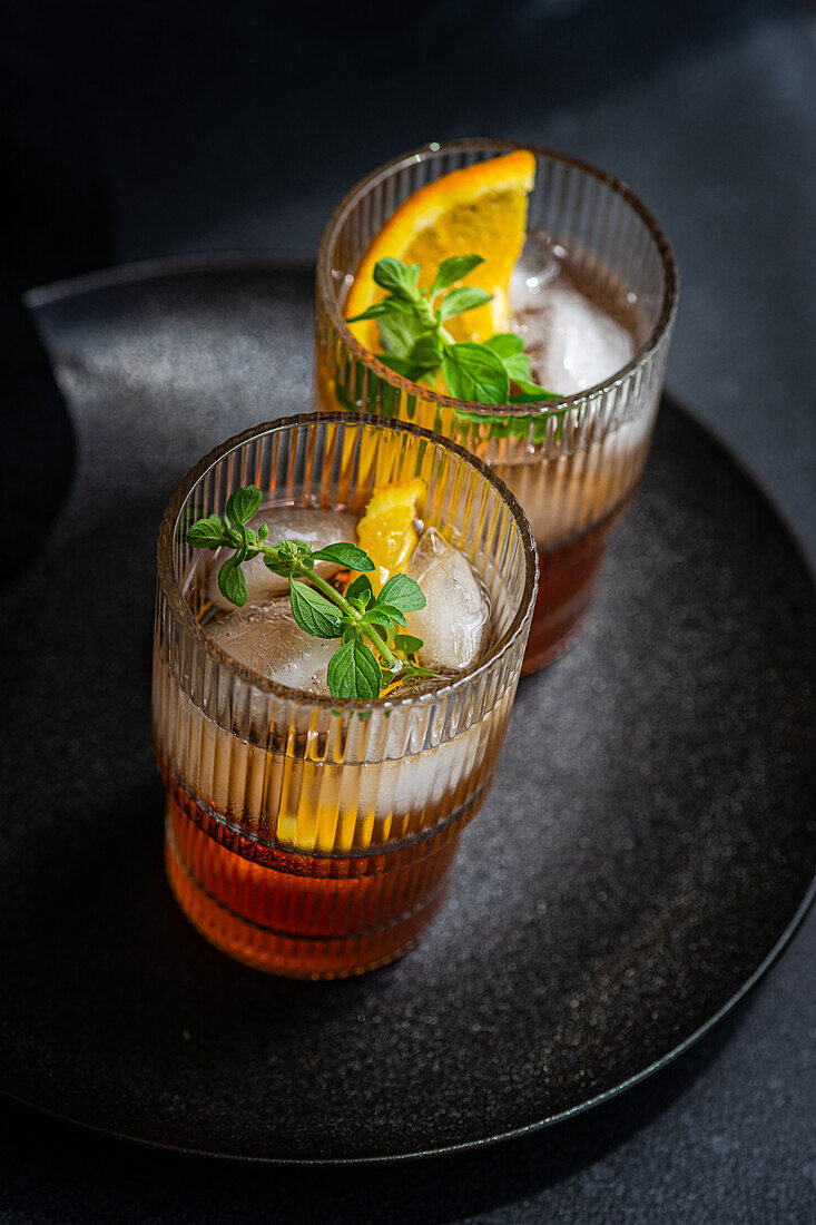 Two elegant glasses filled with cherry liqueur and tonic, garnished with ice, fresh orange slices, and mint leaves, served on a dark, textured surface