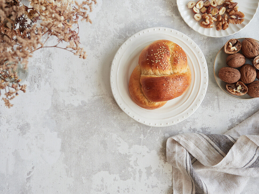 Draufsicht auf eine heitere Herbstmorgenszene mit einem frisch gebackenen Sesambrötchen auf einem Keramikteller, begleitet von einer Auswahl an Walnüssen und Haselnüssen, die einen friedlichen Start in den Tag versprechen