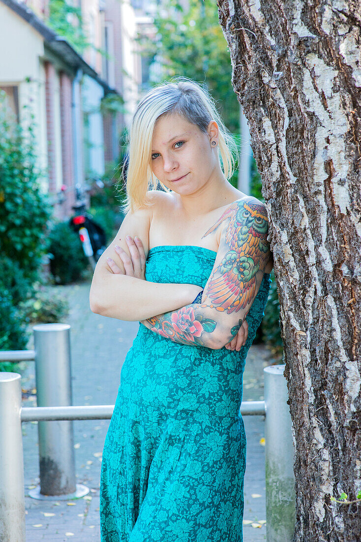 Portrait Young Woman with Tatoos in leaning aginst Tree in Residential Garden. Tilburg, Netherlands.