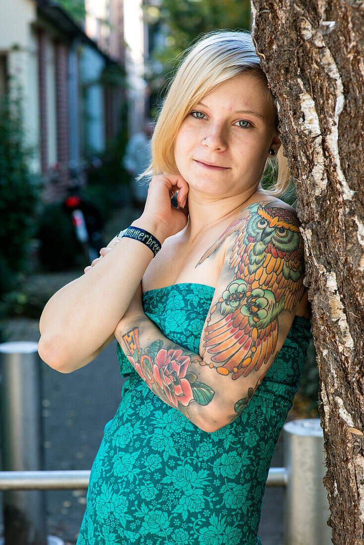 Portrait Young Woman with Tatoos in leaning aginst Tree in Residential Garden. Tilburg, Netherlands.