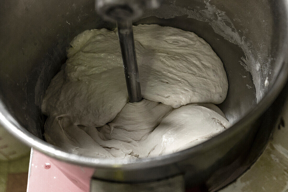 Fresh pizza dough is being thoroughly mixed in a large stainless steel industrial mixer, highlighting the initial stages of pizza preparation