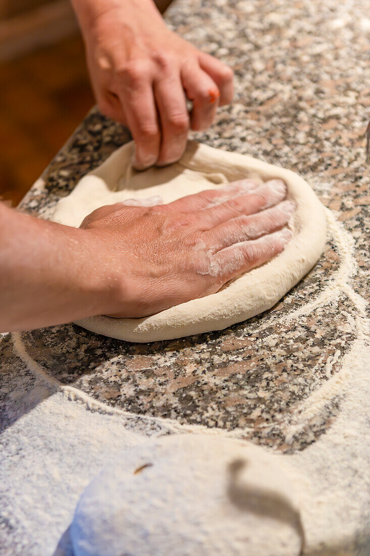 Nahaufnahme von Händen, die frischen Pizzateig auf einer Küchenarbeitsplatte aus Granit formen, die mit Mehl bestreut ist, was den Prozess der Pizzaherstellung verdeutlicht