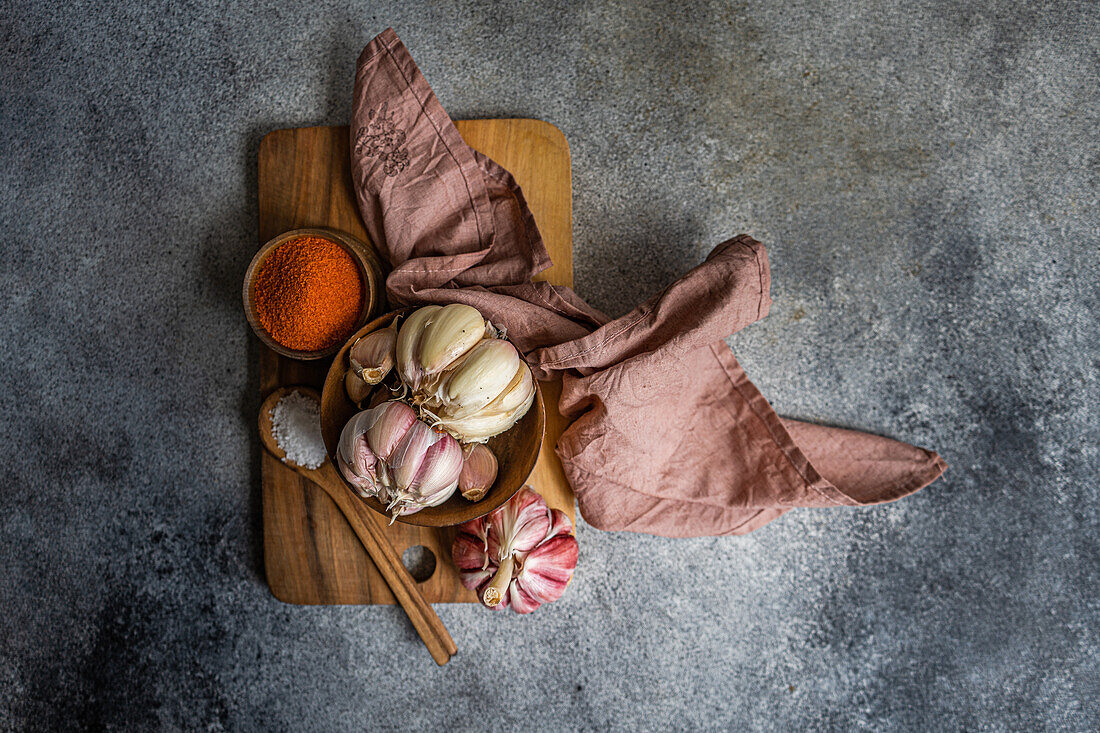 Fresh whole garlic bulbs alongside spices on a wooden cutting board with a textured gray background A linen cloth adds a touch of warmth to the composition