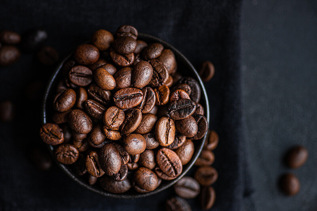 Top view of a dark bowl filled with glossy, fresh coffee beans set against a dark textured background, enhancing the rich textures and deep brown colors.