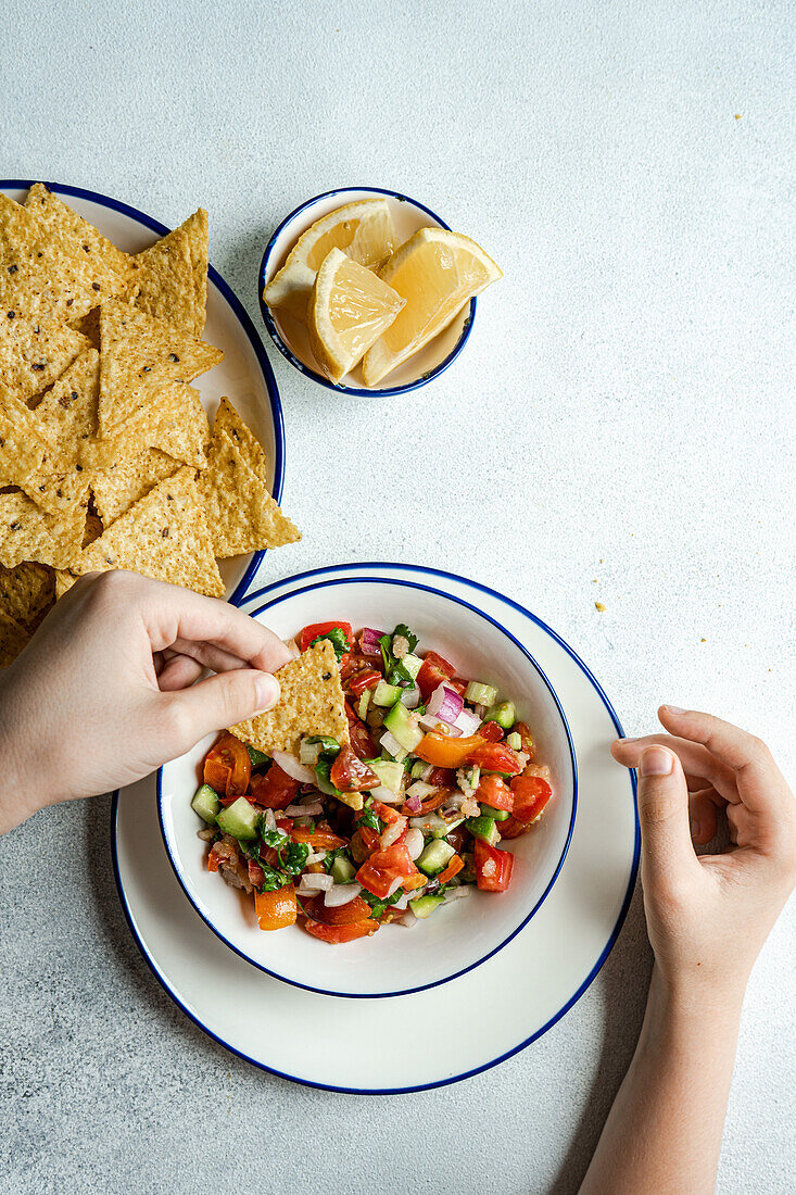 Eine anonyme Person mit einer bunten Schüssel Tomaten-Avocado-Salsa, dekoriert mit gehacktem frischem Koriander und serviert mit knusprigen Maischips und Zitronenscheiben