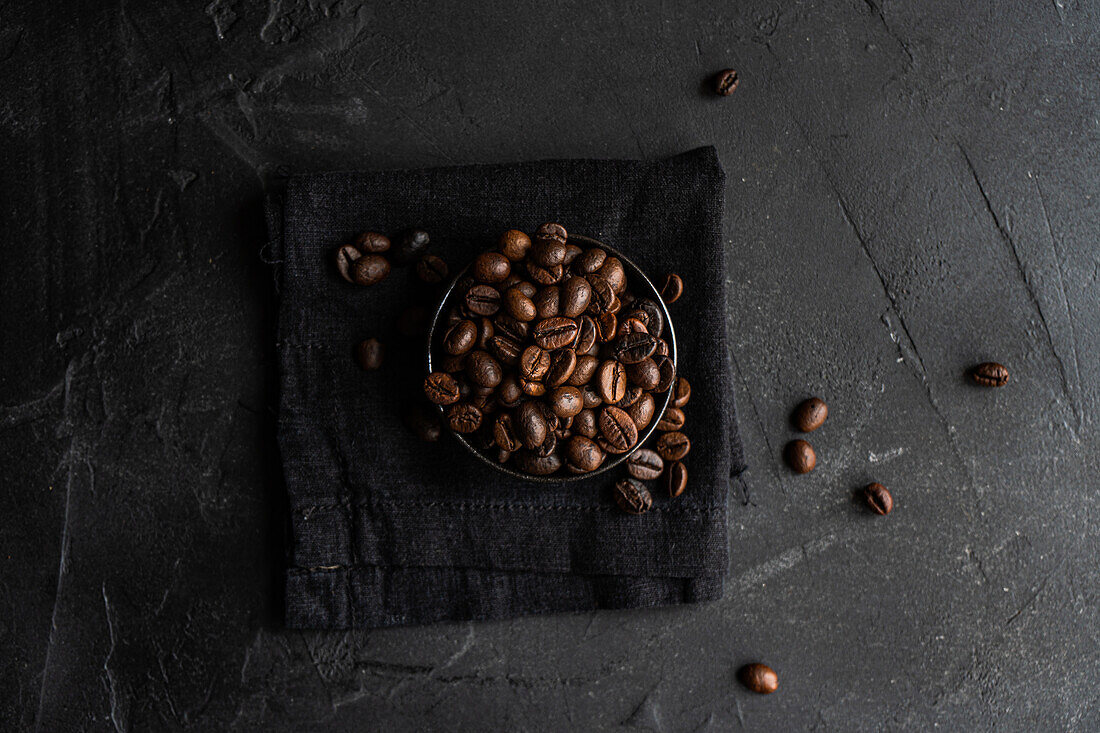 Draufsicht auf dunkel geröstete Kaffeebohnen, die elegant in einer kleinen Metallschale auf einer strukturierten schwarzen Serviette und einer dunkelgrauen, rauen Oberfläche angeordnet sind. Die verstreuten Bohnen verstärken den optischen Reiz und betonen Qualität und Frische.