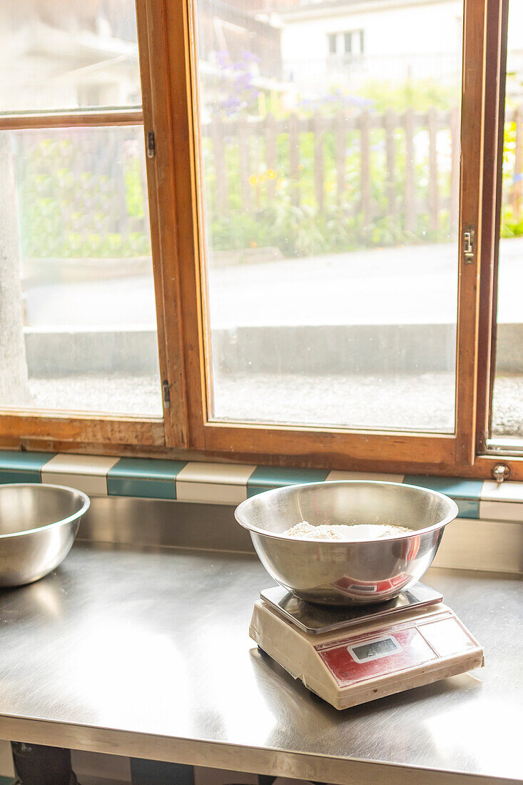 A digital kitchen scale with a bowl of flour on a stainless steel countertop, ready for pizza dough preparation, with a bright window view