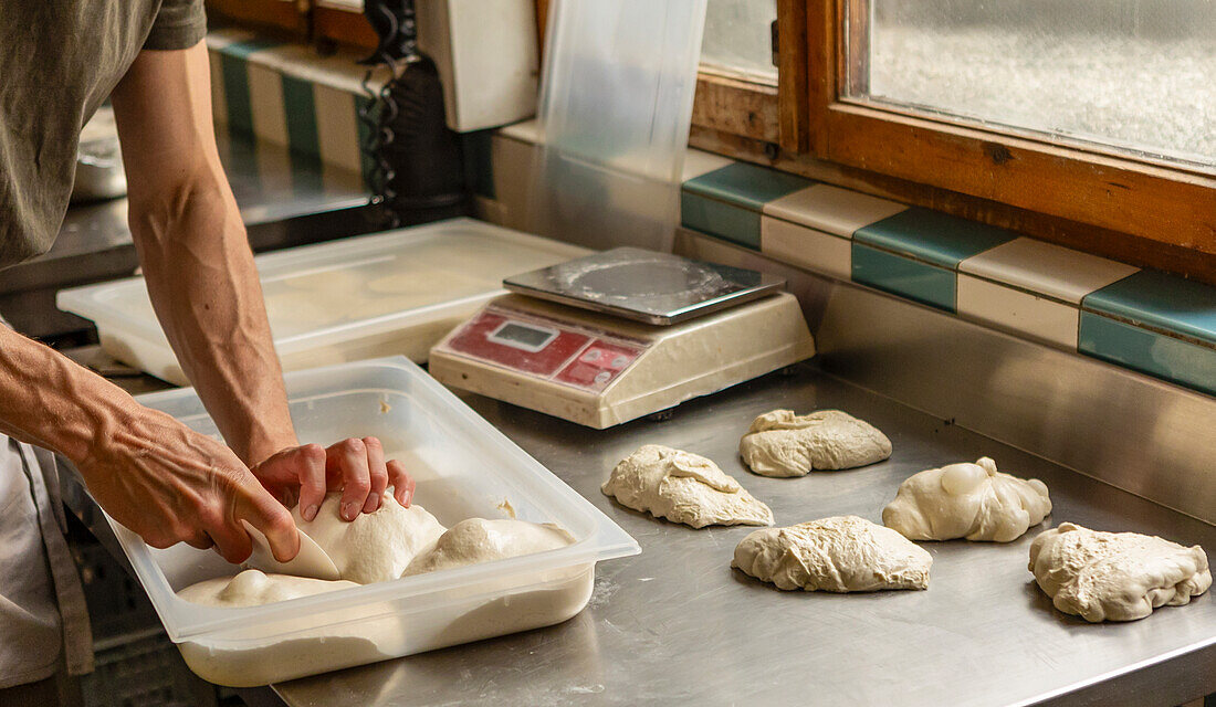 Die geschickten Hände eines Kochs formen akribisch Pizzateigkugeln auf einer Edelstahltheke, eine digitale Waage und ein Vorbereitungstisch sind in einer professionellen Küche zu sehen