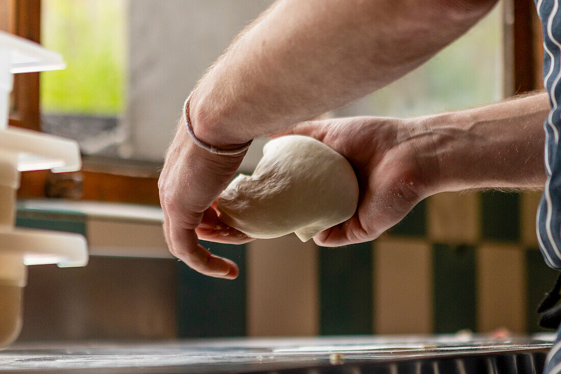 A chef intricately stretches and kneads a ball of pizza dough in a sunlit kitchen, emphasizing the detailed texture and careful process