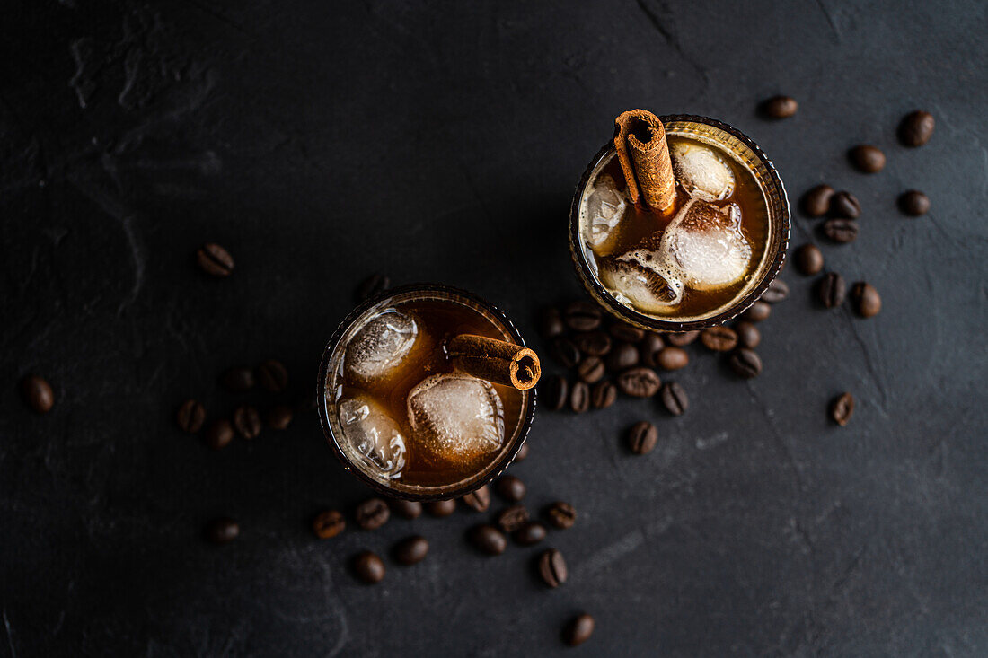 Top view two glasses of chilled coffee adorned with cinnamon sticks and floating ice cubes, set against a backdrop of scattered coffee beans on a dark surface.