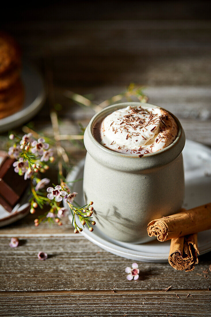 Mexican hot chocolate with whipped cream on a dark wooden background with flowers, biscuits and cinnamon sticks.