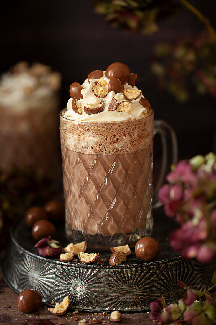 A glass mug with malted hot chocolate topped with cream and chhcolate malt balls.