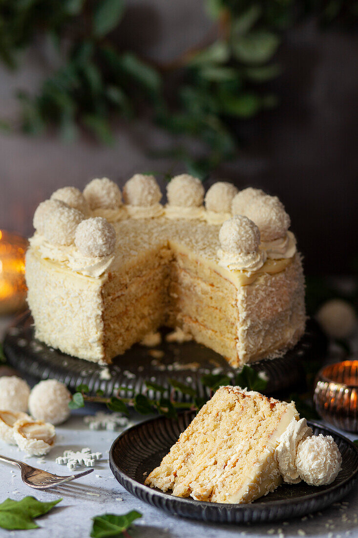 A slice of white chocolate and coconut cake on a plate with the rest of the cake in the background.