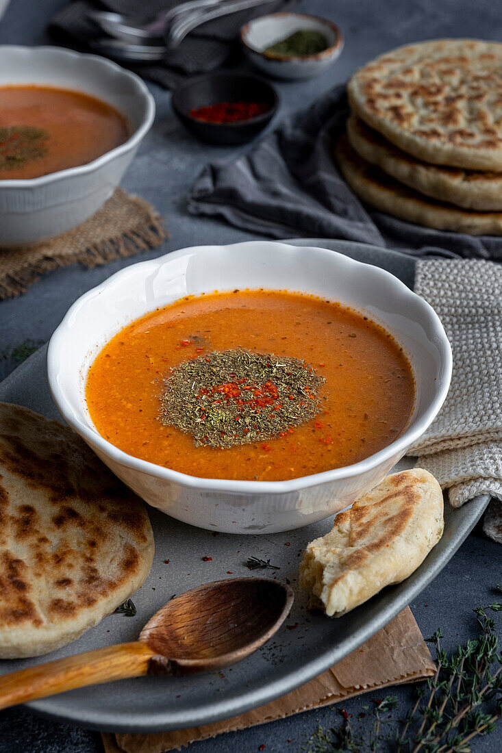 A bowl of tarhana soup, garnished with dried mint and red paprika flakes, some bread and a wooden spoon on the side.