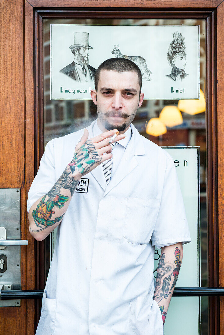 Porträt eines professionellen Friseurs, der seine Zigarette raucht, direkt vor seinem Shorem Barber Shop. Nieuwe Binnenweg, Rotterdam, Niederlande.