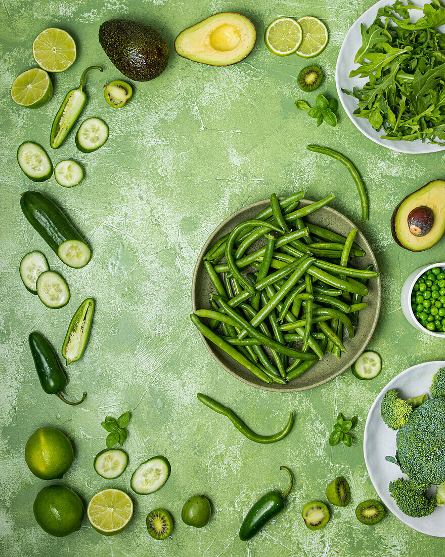 Green vegetables on a green backdrop, including peas, avocado, cucumber, beans, brocolli, tomatoes and peppers