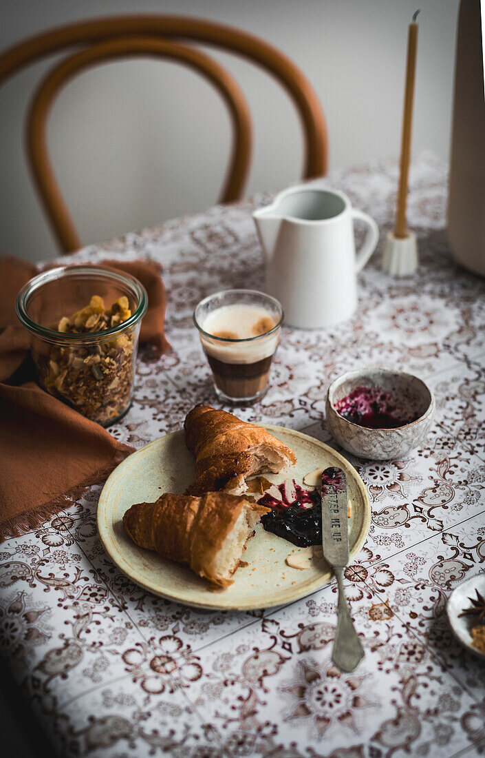 A breakfast scene, a rustic chair and a table with an espresso, a croissant and blueberry jam and muesli.